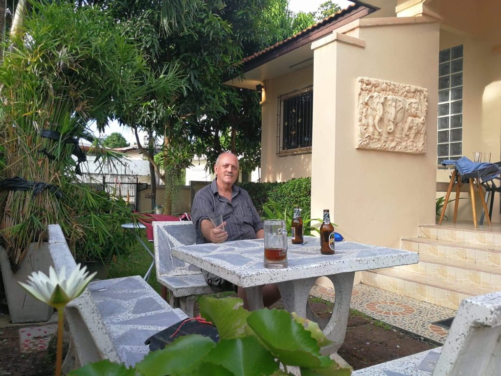 Man sitting at a stone outdoor table with beer bottles, in a sunny garden next to a beige house with a carved wall relief.