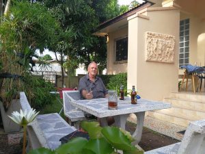 Man sitting at a stone outdoor table with beer bottles, in a sunny garden next to a beige house with a carved wall relief.