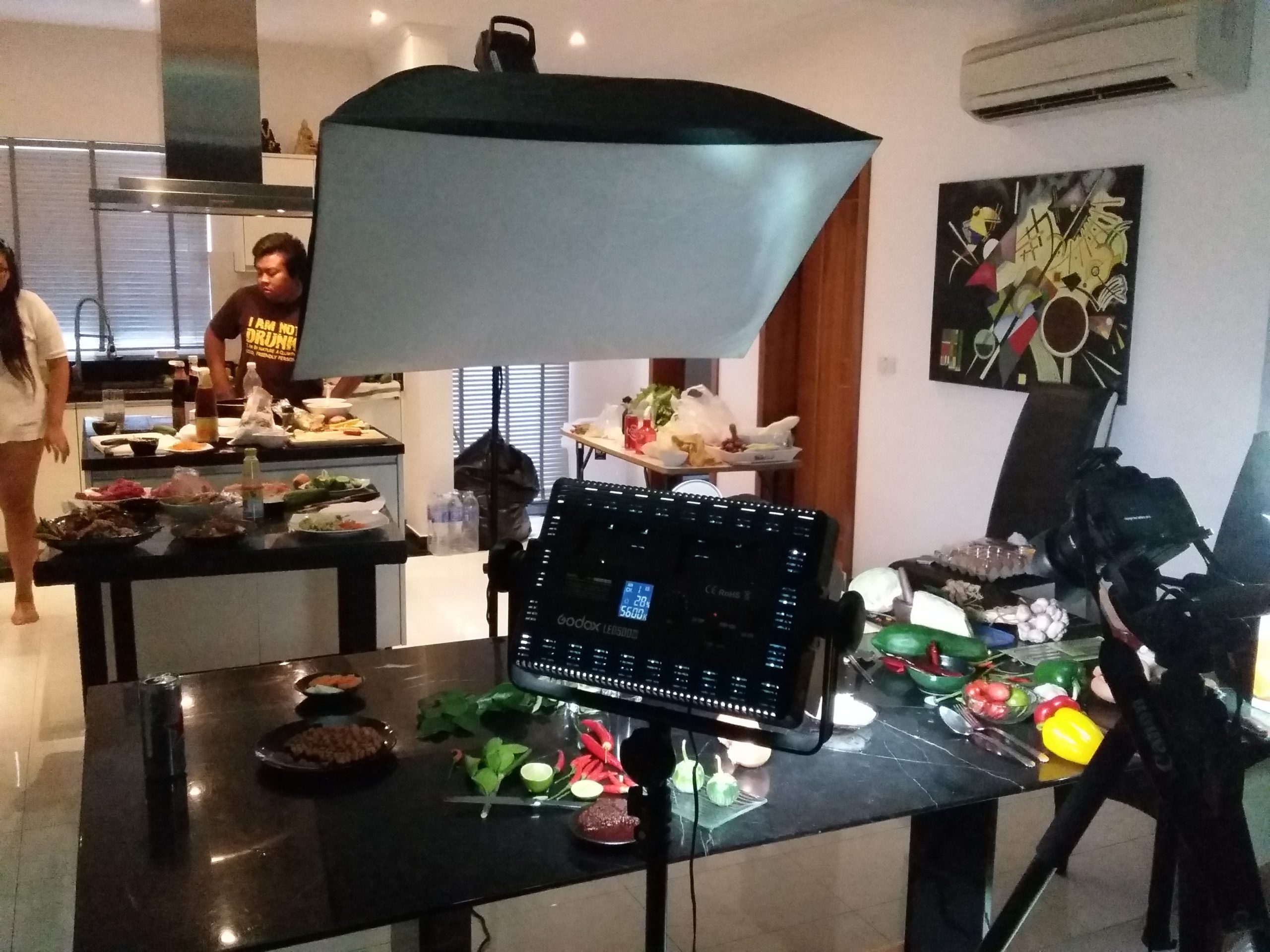 Large studio light hangs over a kitchen island filled with platters of food and drinks, with people in the background.